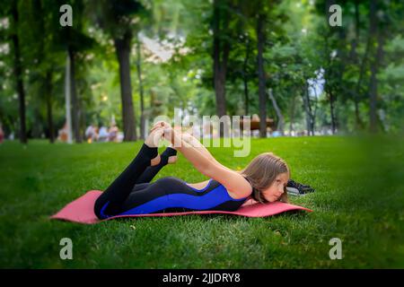 nel parco, una bambina si piega indietro, si aggrappa le gambe, esegue gli elementi yoga, si allunga Foto Stock