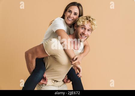 Coppia bianca con magliette sorridenti mentre piggybacking isolato su sfondo beige Foto Stock