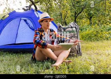 Defocus giovane donna che lavora sul tablet vicino campeggio tenda all'aperto circondato da bella natura. Freelance, sabbatico, salute mentale. Direzione della mano. Foto Stock