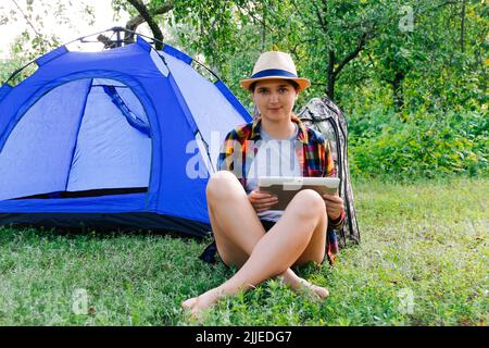 Defocus giovane donna che lavora sul tablet vicino campeggio tenda all'aperto circondato da bella natura. Freelance. Fuori fuoco. Foto Stock