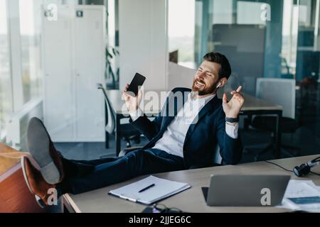 Un uomo d'affari allegro che si rilassa in ufficio, si riposa sul tavolo e si diverta con la musica nelle cuffie, prendendo pausa dal lavoro Foto Stock