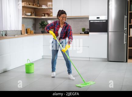 Allegra casalinga nera millenaria in guanti di gomma con mop lava il pavimento, gode di pulizia Foto Stock