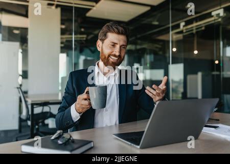 Bell'uomo d'affari caucasico positivo in tuta seduta in ufficio, parlando con i partner commerciali via laptop Foto Stock