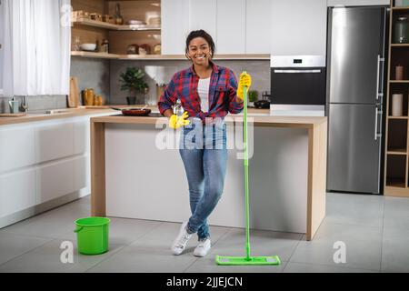 Sorridente donna americana africana casalinga in guanti di gomma con mop lava pavimento, gode di pulizia Foto Stock