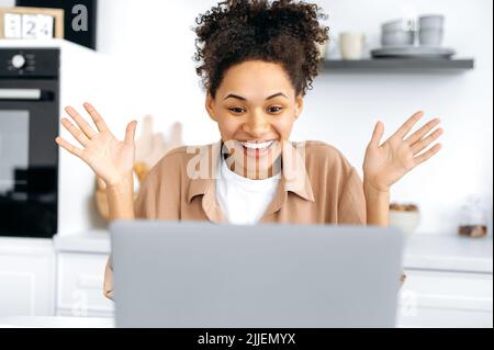 Allegro gioioso giovane afroamericana, seduto in cucina a casa, usando il suo laptop, eccitato guardando lo schermo, facendo il gesto vincitore con le braccia, celebrando il successo, sorridendo felicemente Foto Stock