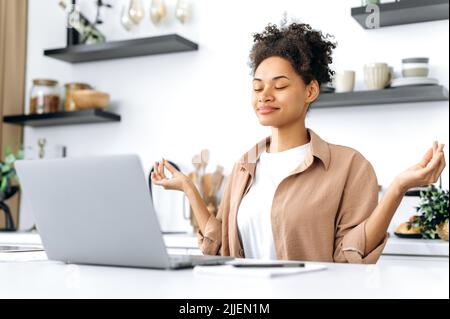 Calmo afroamericano giovane donna, studente o freelance, si siede in cucina al tavolo, prende una pausa dal lavoro o dallo studio, medita, rilassa il cervello, sorridendo. Meditazione e relax Foto Stock