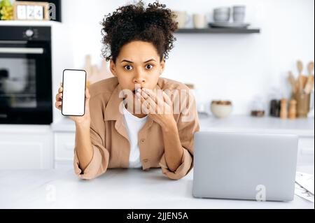 Scioccata giovane afroamericana eccitato, si alza in cucina vicino al tavolo con un laptop, tiene uno smartphone in mano con bianco bianco bianco mock-up schermo, stupito guarda la macchina fotografica, coprendo la sua bocca Foto Stock