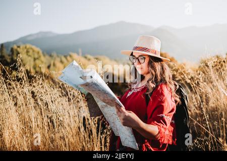 Donna viaggiatore latino che indossa un cappello, un poncho rosso e uno zaino, controllando la mappa durante il trekking per esplorare la natura. Foto Stock