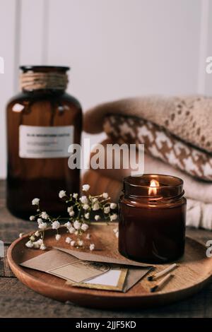 Candela che brucia in un piccolo vaso di vetro ambrato, fiori di gypsofila e maglioni di condimenti a maglia impilati. Stile di vita accogliente, concetto hygge Foto Stock