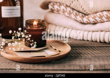 Candela che brucia in un piccolo vaso di vetro ambrato, fiori di gypsofila e maglioni di condimenti a maglia impilati. Stile di vita accogliente, concetto hygge Foto Stock