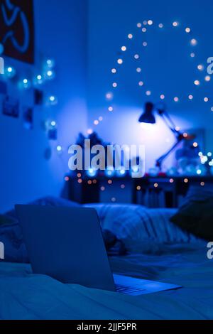 Primo piano di un computer portatile sul letto in una stanza buia decorata con luci al neon blu in una stanza adolescente Foto Stock