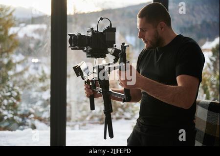 Uomo videografo che realizza riprese, utilizzando una fotocamera montata su un'attrezzatura stabilizzatore gimbal. Finestre panoramiche con paesaggio invernale sullo sfondo. Foto Stock