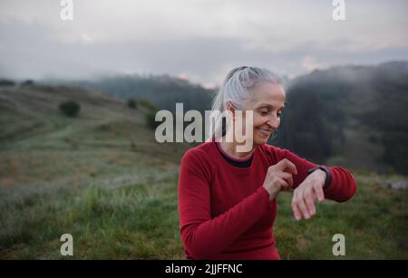 Jogger donna anziana impostazione e guardare lo smartwatch sportivo, controllando le sue prestazioni in natura al mattino presto con nebbia e montagne sullo sfondo Foto Stock