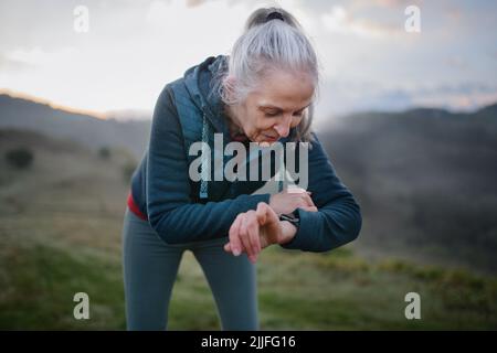 Jogger donna anziana impostazione e guardare lo smartwatch sportivo, controllando le sue prestazioni in natura al mattino presto con nebbia e montagne sullo sfondo Foto Stock