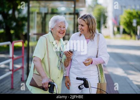 Ritratto di caregiver con donna anziana a piedi nel parco con shopping bag, ridendo e parlando. Foto Stock