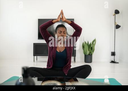 Primo piano di donna africana nera seduta in posa yoga sul tappeto in soggiorno, stretching e meditazione a casa di allenamento Foto Stock