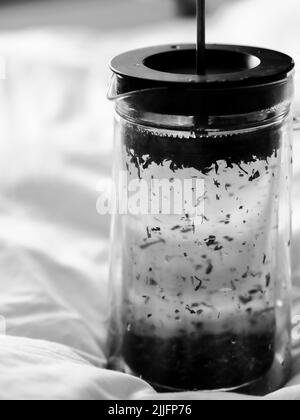 Uno scatto verticale in scala di grigi di un French Press con tè su lenzuola Foto Stock