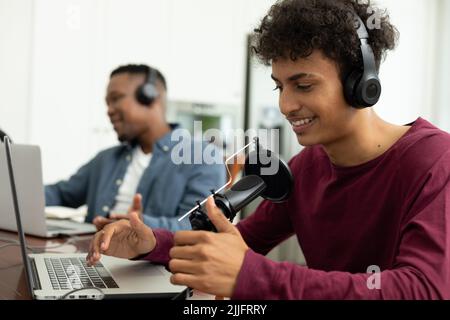 Sorridente podcaster maschile multirazziale che parla al microfono mentre registra podcast con un collega Foto Stock