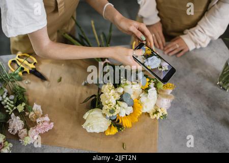 Mani di due giovani fiorista che lavorano in un negozio di fiori. Fiorista femminile allegro scattare foto di piante in negozio floreale utilizzando la fotocamera del telefono cellulare. Primo piano. Foto Stock