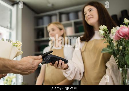 Due fioristi sorridenti che tengono la macchina del lettore di carte al banco mentre il cliente paga con la carta di credito elettronica nel suo smartphone. Foto Stock