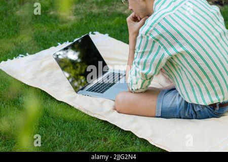 Vista posteriore del giovane uomo senza volto concentrato utilizzando il computer seduto all'aperto sotto un albero in primavera Foto Stock