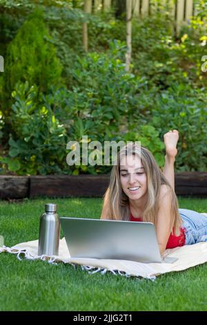 Bella giovane donna in abito rosso che guarda la macchina fotografica sorridente e felice mentre usando il suo laptop circondato dalla natura in primavera Foto Stock