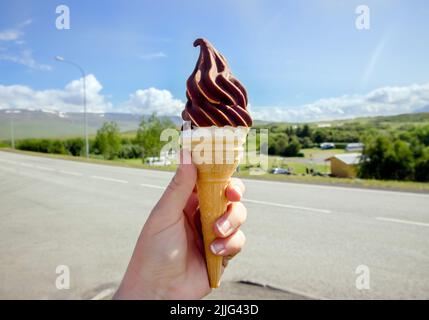 Tenuta di mano gustoso islandese locale soft serve gelato alla vaniglia immerso in cioccolata calda, Islanda natura sullo sfondo. Foto Stock