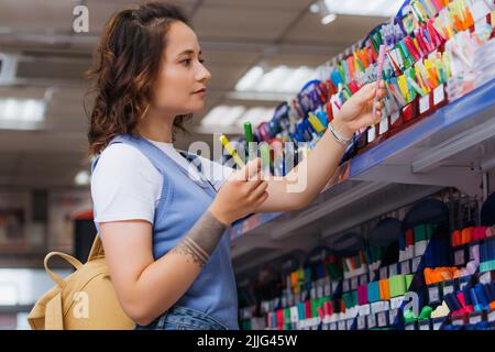 vista laterale della donna tatuata che tiene penne multicolore vicino a rack in negozio Foto Stock