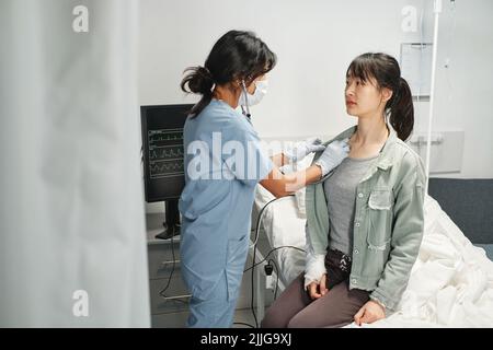 Operatore medico che indossa maschera protettiva sul viso utilizzando stetoscopio ascoltare i suoni interni della giovane donna asiatica Foto Stock