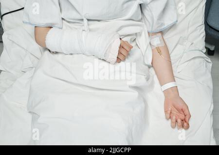 Shot orizzontale dall'alto verso il basso di una paziente femminile non riconoscibile con braccio rotto che riceve farmaci o fluidi nutrienti attraverso la linea endovenosa Foto Stock
