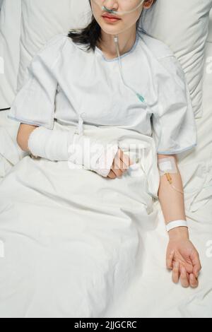 Shot verticale ad angolo alto di giovane paziente asiatico con braccio rotto in colata che riceve farmaci o fluidi nutrienti attraverso la linea endovenosa Foto Stock