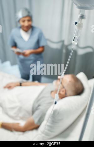 Fuoco selettivo sul primo piano shot dell'uomo maturo che ottiene la terapia IV nell'ospedale moderno che parla al suo medico Foto Stock