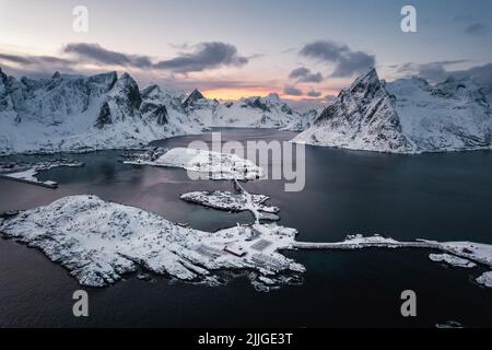Scatto con drone aereo del bellissimo villaggio di pescatori di Reine nelle isole Lofoten durante l'inverno al tramonto circondato da montagne innevate. Foto Stock