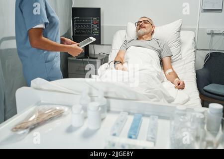 Un operatore medico irriconoscibile che tiene un tablet digitale che parla con l'uomo maturo che controlla il suo stato di salute nella stanza dell'ospedale Foto Stock