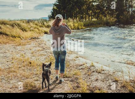 vista posteriore della giovane donna che cammina lungo la riva del lago con cani di razza mista viaggio ed escursioni con animali domestici Foto Stock