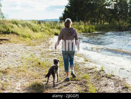 vista posteriore della giovane donna che cammina lungo la riva del lago con cani di razza mista viaggio ed escursioni con animali domestici Foto Stock