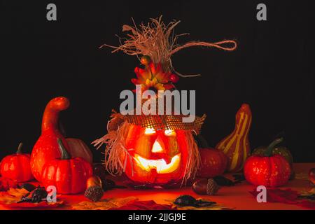 Una zucca illuminata di Halloween adornata con un cappello e fiori circondati da foglie, ghiande, zucche e ragni su una superficie arancione con una b nera Foto Stock