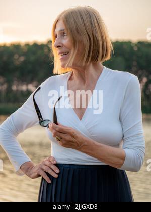 gioiosa senior fit elegante donna vestita bianco e blu indossando occhiali da sole godendo la natura in estate al tramonto Foto Stock