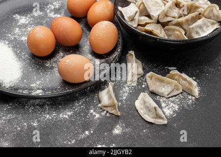 Uova sul piatto nero. Gnocchi di gyoza crudi in ciotola e sul tavolo. Vista dall'alto. Sfondo nero Foto Stock