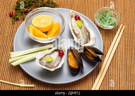 Vongole e ostriche in conchiglie, limone in ciotola e gambi di sedano in ciotola grigia. Chopsticks, alghe in ciotola e rametti di timo su tovagliolo in legno sottile Foto Stock