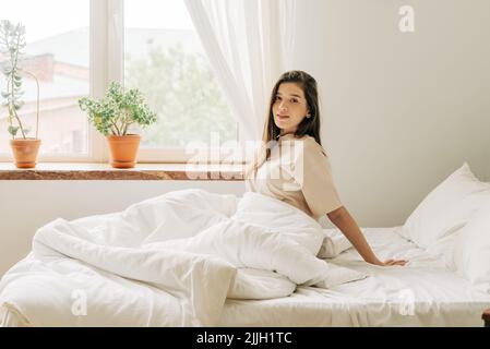 Giovane e attraente brunette si siede sul letto accanto alla finestra dopo il risveglio al mattino. Foto Stock