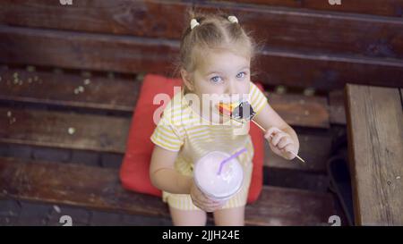 la ragazza della tartaruga mangia un pan di zenzero colorato e tiene un milkshake nella sua mano. Primo piano di ragazza carina bambino seduto sul panca del parco e mangiare biscotti con un Foto Stock