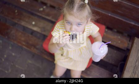 la ragazza della tartaruga mangia un pan di zenzero colorato e tiene un milkshake nella sua mano. Primo piano di ragazza carina bambino seduto sul panca del parco e mangiare biscotti con un Foto Stock