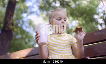 la ragazza della tartaruga mangia un pan di zenzero colorato e tiene un milkshake nella sua mano. Primo piano di ragazza carina bambino seduto sul panca del parco e mangiare biscotti con un Foto Stock