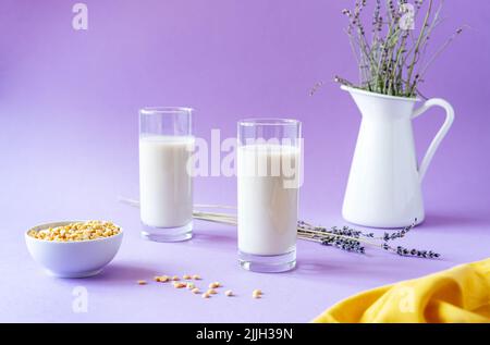 Pisello vegano o latte di lavanda a base di piante in bicchieri, semi di piselli, caraffa bianca con lavanda, tovagliolo giallo. Sfondo lilla. Spazio di copia Foto Stock