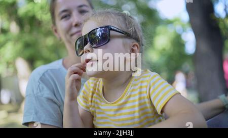 Primo piano di mamma mette i suoi occhiali da sole su carina bambina seduta nel parco. Primo piano di madre e figlia giocando mentre si siede su una panca del parco in un Foto Stock