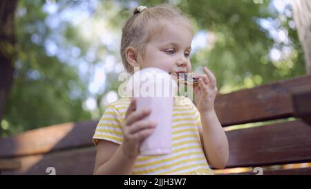 Odessa, Ucraina, Europa dell'Est. 26th luglio 2022. La ragazza della tartaruga mangia un pan di zenzero colorato e tiene un milkshake in mano. Primo piano di ragazza carina bambino seduto sul panchina del parco e mangiare biscotti con un milkshake. (Credit Image: © Andrey Nekrasov/ZUMA Press Wire) Foto Stock