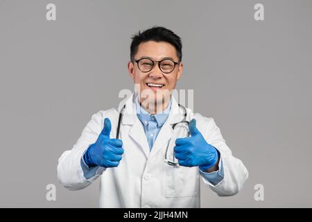 Felice uomo medico giapponese di mezza età in cappotto bianco, occhiali e guanti protettivi mostra pollici in su Foto Stock