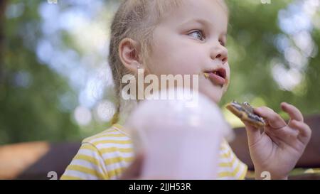 Odessa, Ucraina, Europa dell'Est. 26th luglio 2022. La ragazza della tartaruga mangia un pan di zenzero colorato e tiene un milkshake in mano. Primo piano di ragazza carina bambino seduto sul panchina del parco e mangiare biscotti con un milkshake. (Credit Image: © Andrey Nekrasov/ZUMA Press Wire) Foto Stock