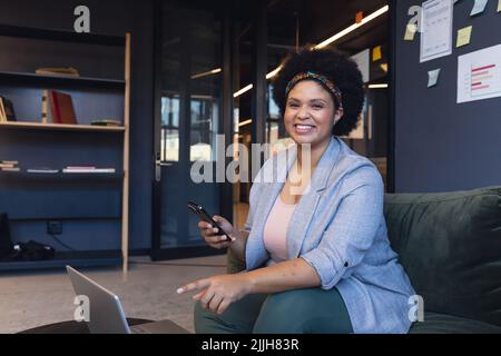 Ritratto di donna d'affari biraciale sorridente con smartphone seduto in ufficio Foto Stock